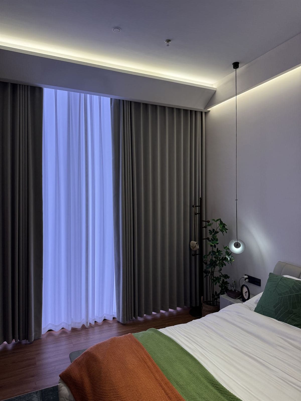 A bedroom at dusk with motorised sheer and blackout curtains, warm cove LED along the ceiling, and a small GUOCIO pendant beside the bed