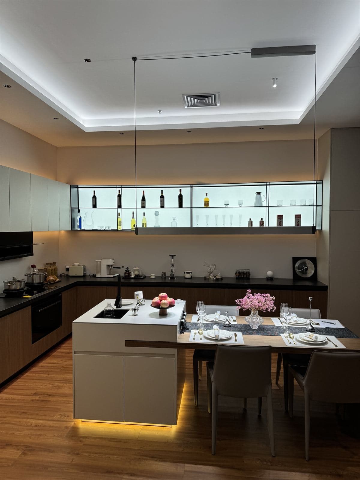 An open-plan kitchen and dining room layered with warm cove lighting, an illuminated glass display shelf and a linear track light over the island