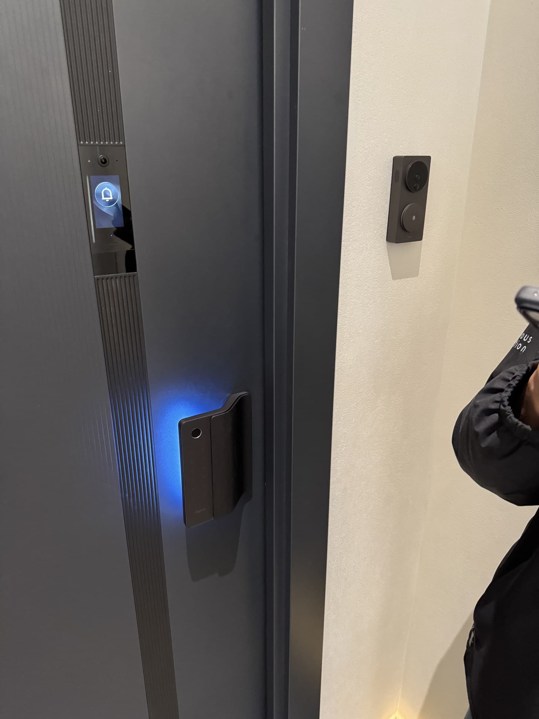 A smart door with a glowing blue handle and a doorbell display showing a bell icon. A smart camera is mounted on the adjacent wall.